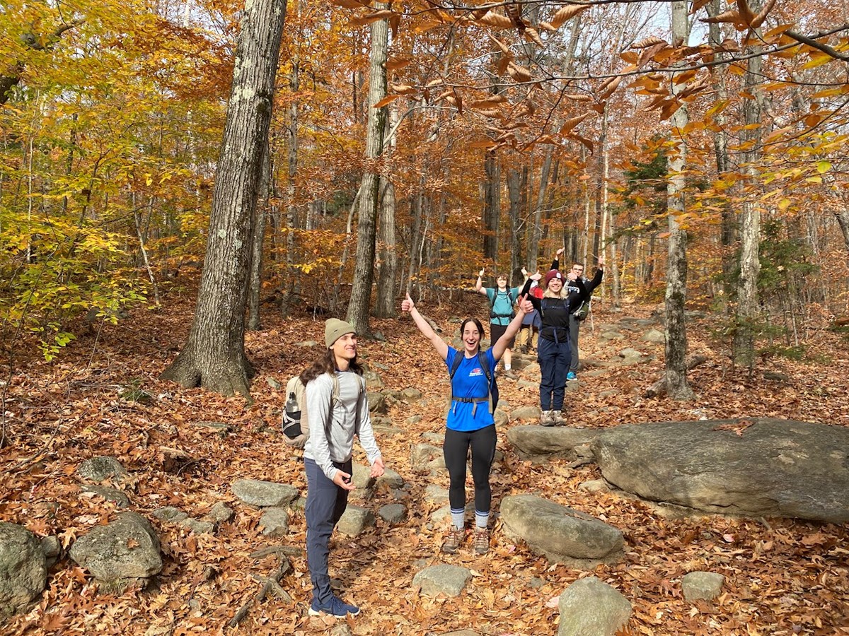 A group smiles with arms in the air on a leaf covered trail in fall.