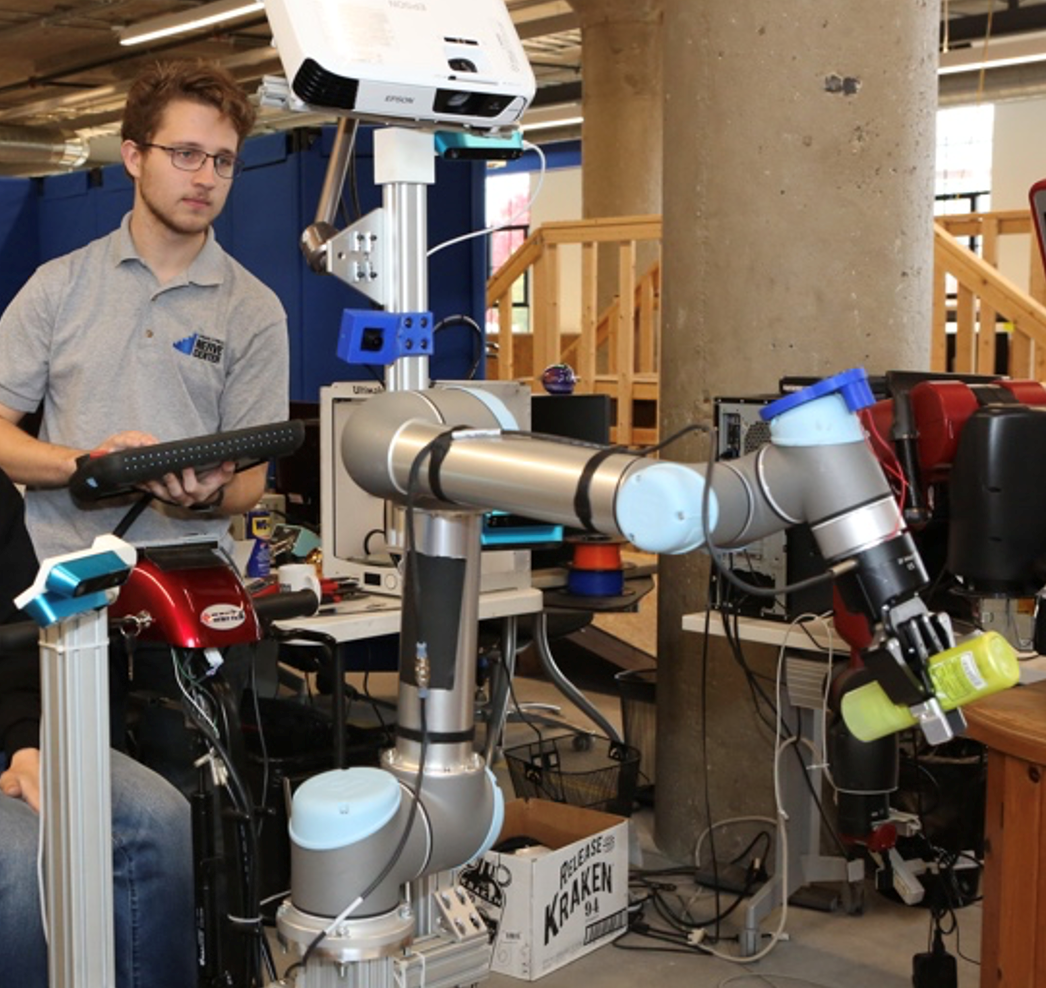A student uses a teach pendant to interact with a robot arm grabbing a bottle.