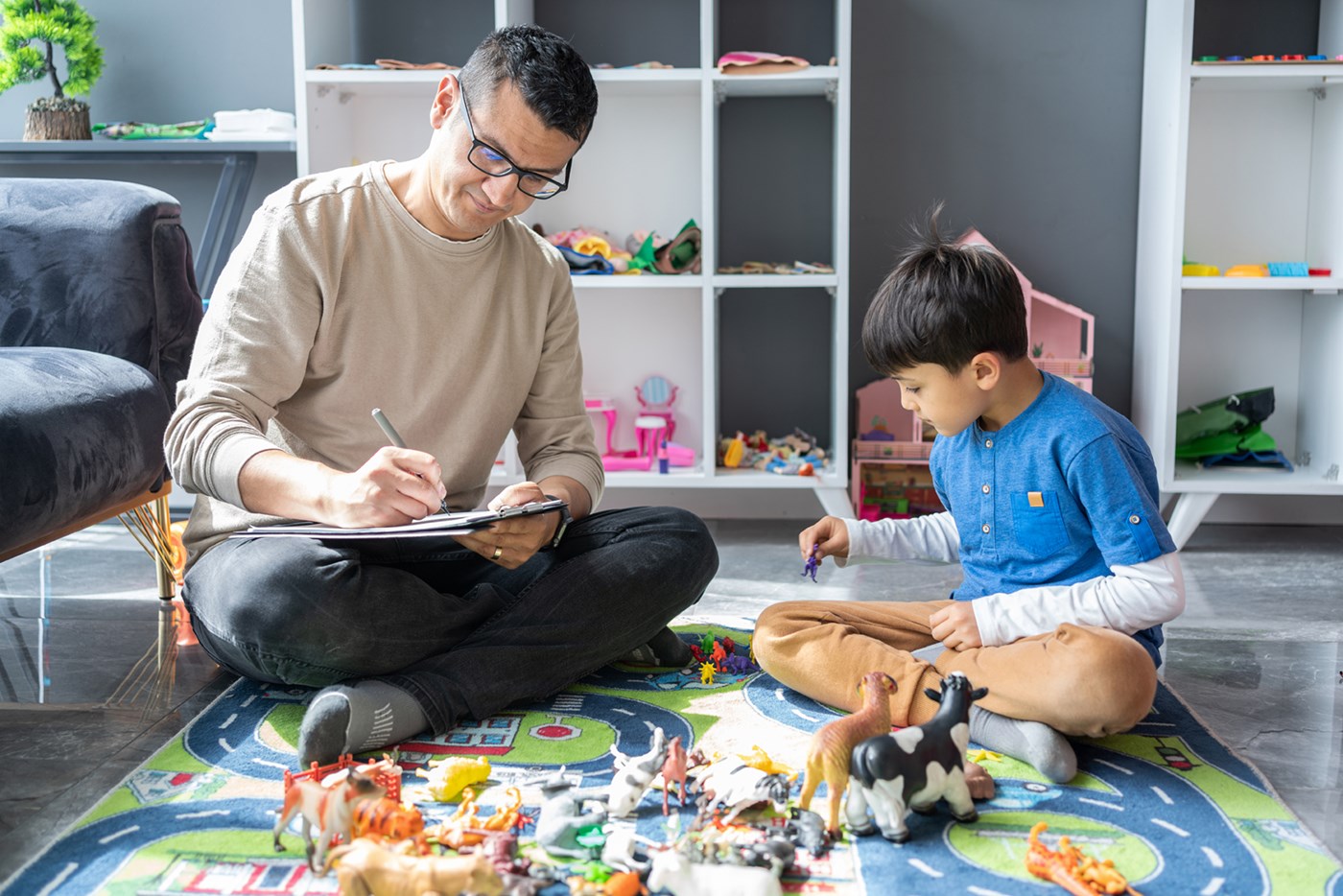 An adult takes notes while sitting next to a child on a floor covered with toys.