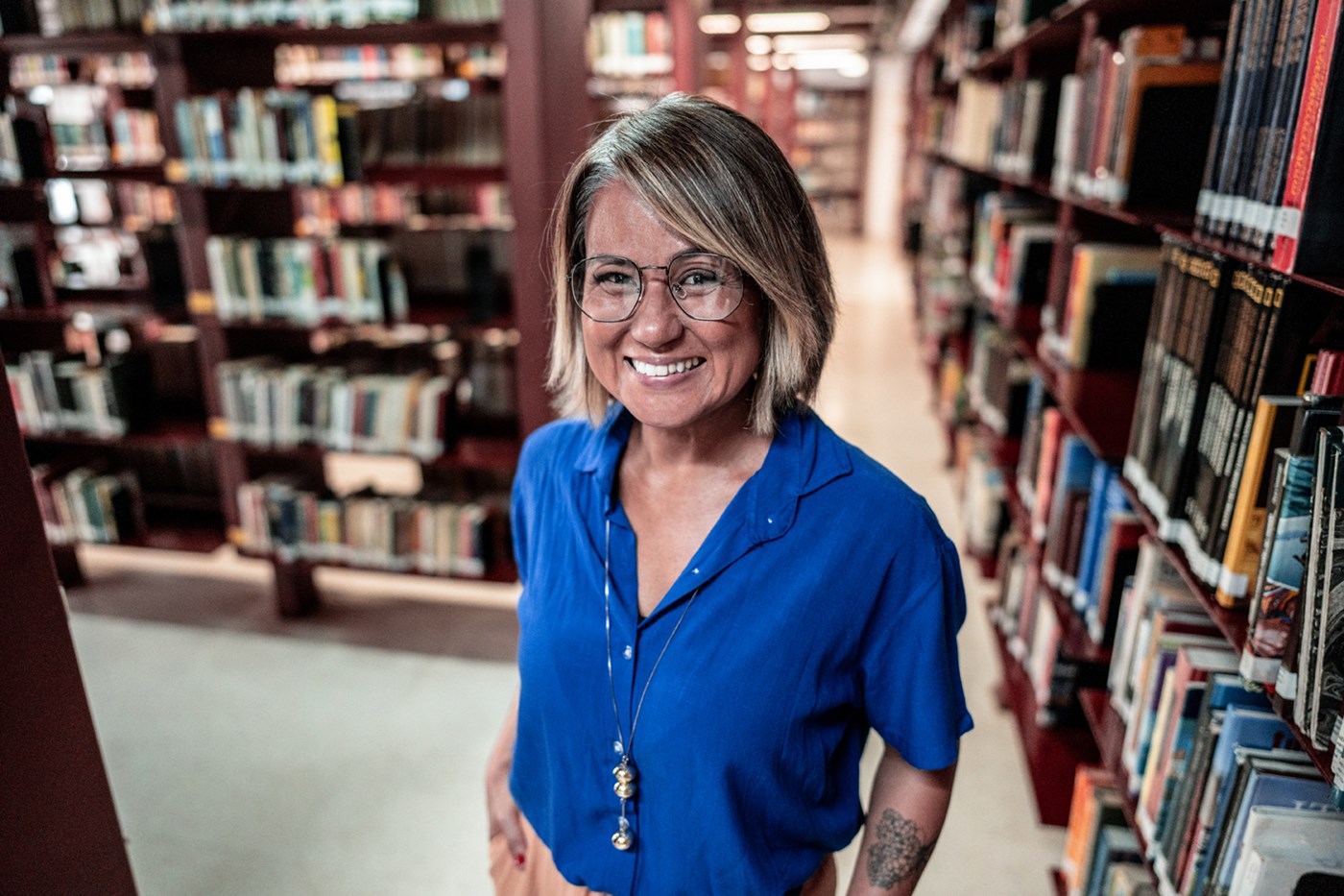 A person smiles while standing among bookshelves in a library.