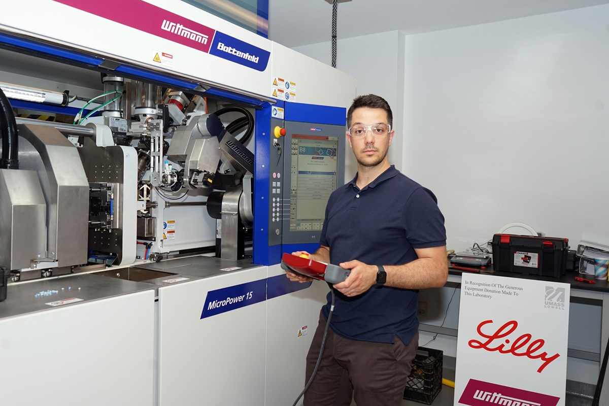 Graduate student Davide Masato standing beside an industrial machine, holding a handheld control device.