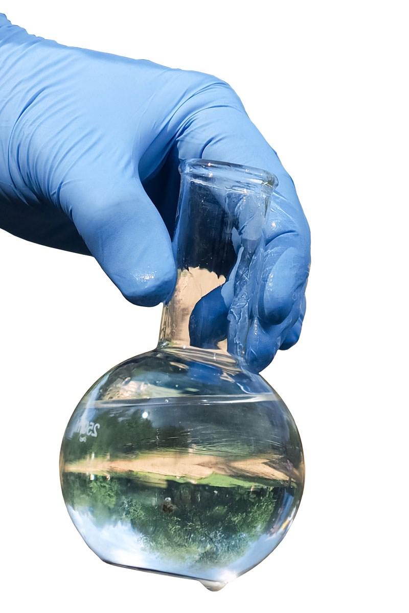 A gloved hand holding a glass flask filled with clear liquid that reflects a green scenery.