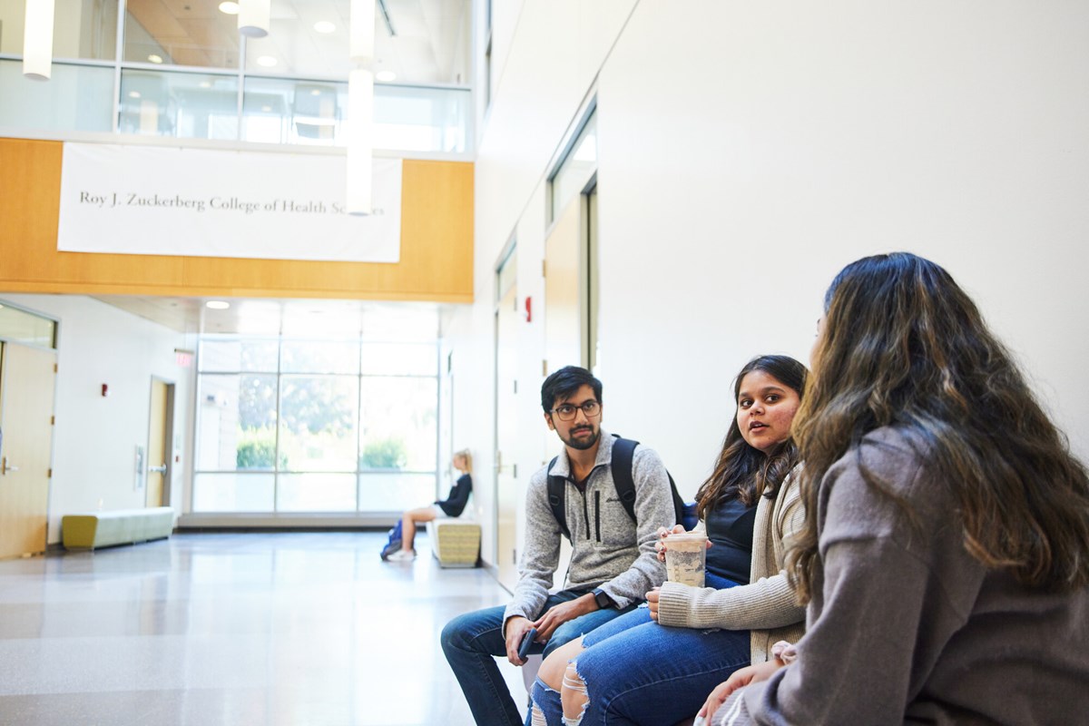 Three students seated on bench chat in hallway of UMass Lowell Health Sciences building