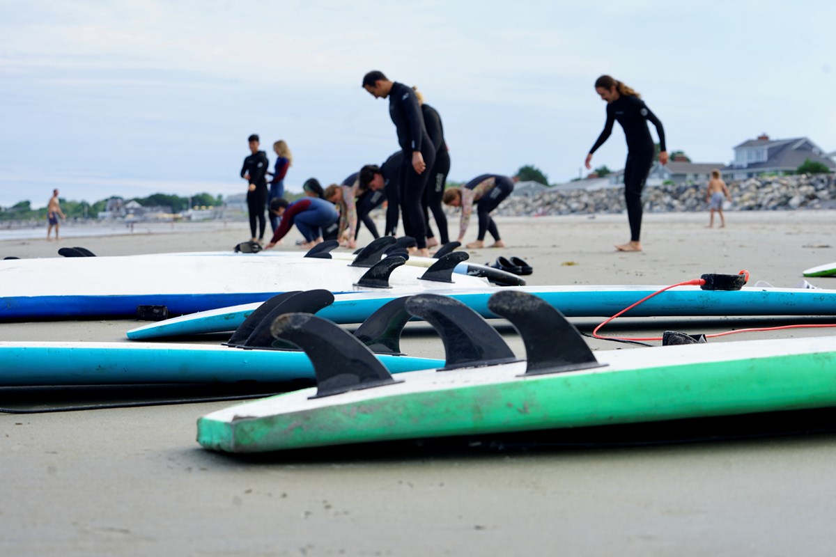 A group of people in wetsuits practice surfing on a beach. There are several surfboards laying in the foreground.