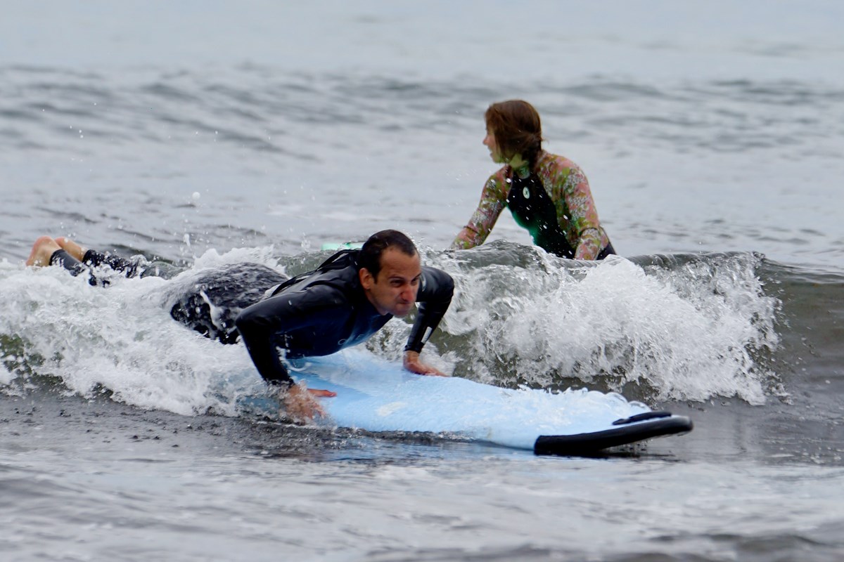 A man in a wetsuit lays on a surfboard catching a wave while a person waits for a wave behind him.