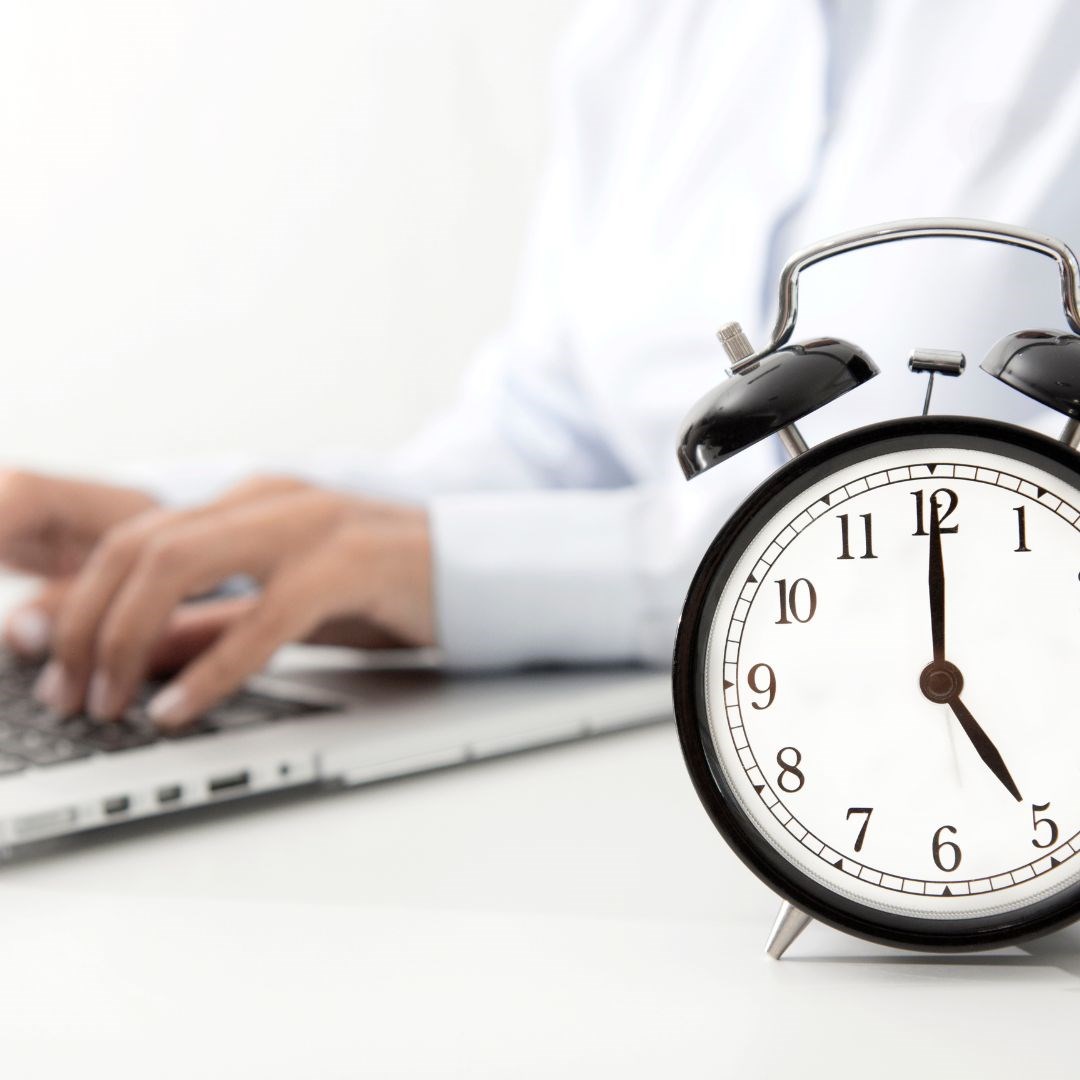 Person typing on laptop next to alarm clock.