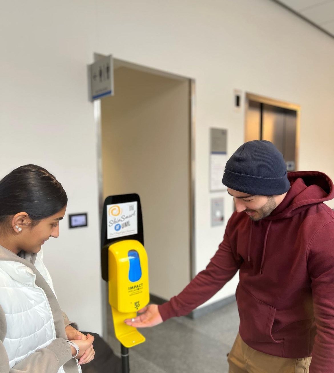 A person shows another how to use an IMPACT melanoma SPF 30 sunscreen dispenser. A sign on the dispenser reads: Skin Smart @ UML.