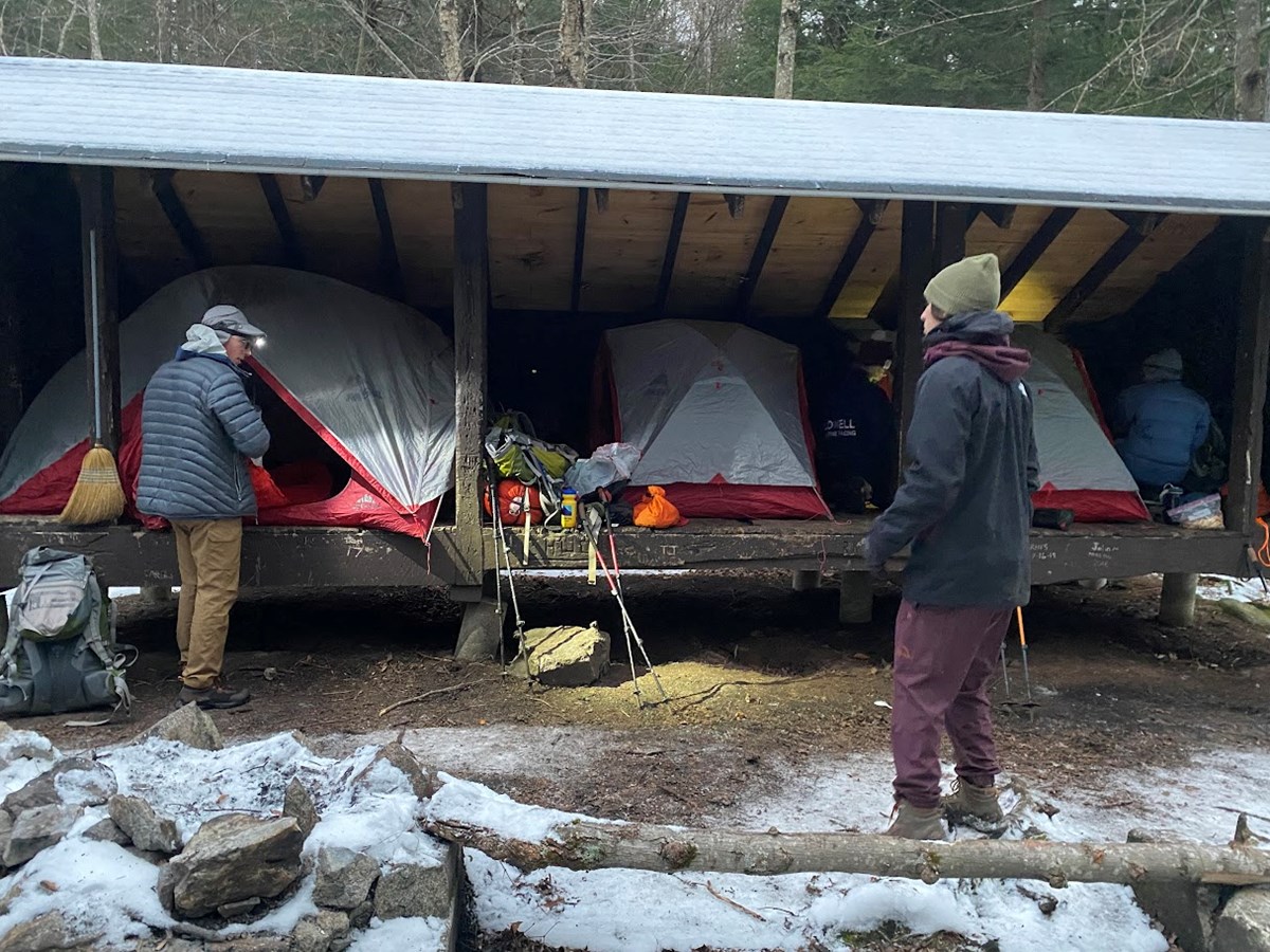 Two people wearing winter gear stand in front of an open-sided wooden shelter with three tents erected inside with a dusting of snow on the ground.