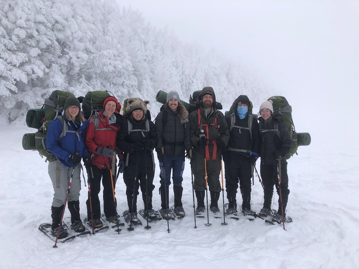 a group of seven stands bundled up carrying backpacks in a snow covered and foggy landscape